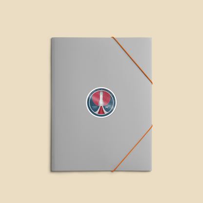 A gray notebook or journal with an orange elastic closure band and a circular red, white, and blue peace sticker in the center, shown on a solid beige background.