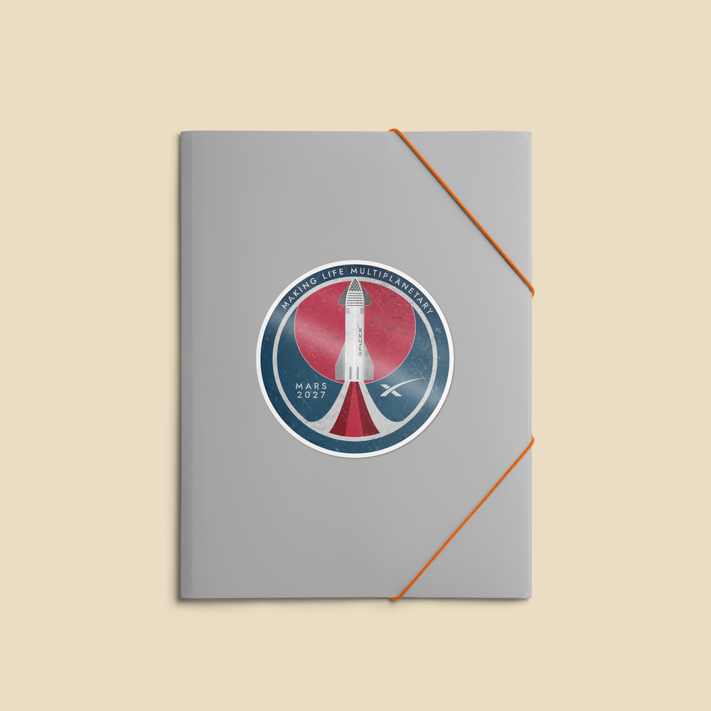 A gray notebook with an orange elastic closure strap, featuring a circular 'Mars 2027 Space Launch' commemorative sticker on the front cover, set against a solid beige background.