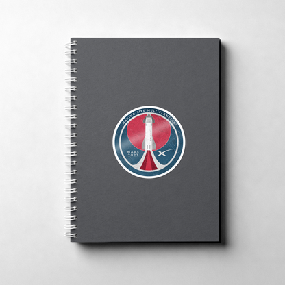 A dark gray spiral notebook with a circular space-themed sticker on the front cover, photographed on a light gray background.