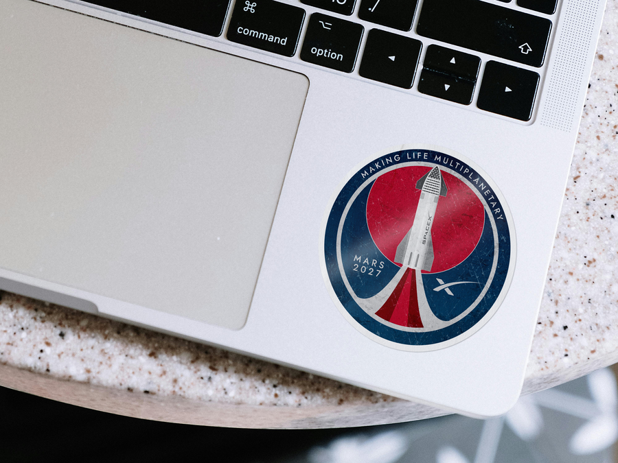 A circular sticker featuring a retro-style rocket launch graphic with the text 'MAKING LIFE MULTIMENSIONAL' and 'MARS 2027', applied to the palm rest of a silver laptop on a speckled countertop.